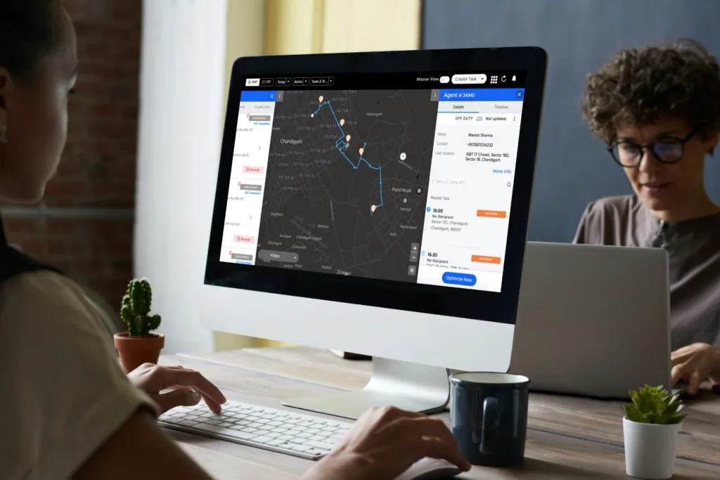 Two people working at desks, one looking at a monitor that shows a dark dispatch map with routes and a job list