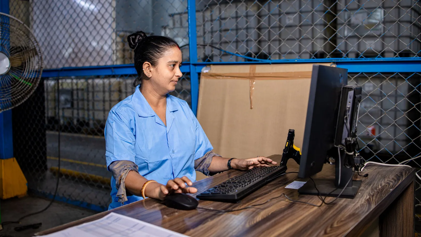 An office worker in a blue work shirt typing at a computer on a desk inside a warehouse, behind a chain-link partition, the kind of office user many field service plans bill as a full user