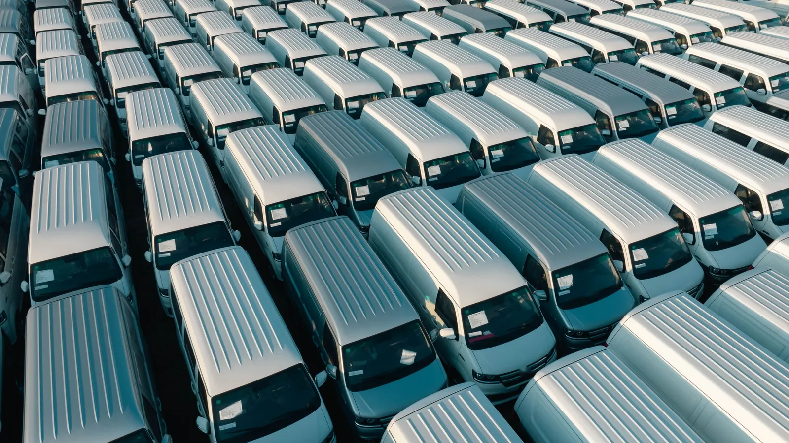 Aerial view of dozens of identical white vans parked in tight rows on a lot, each one a vehicle a per-vehicle GPS add-on would bill every month