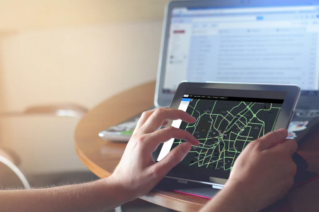 Hands on a tablet showing a city map with route lines, with a laptop in the background